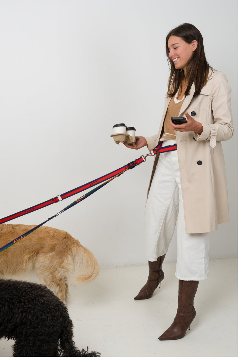 Woman in a trench coat walking two dogs hands free tangle free using the Allez dog leash against a white background